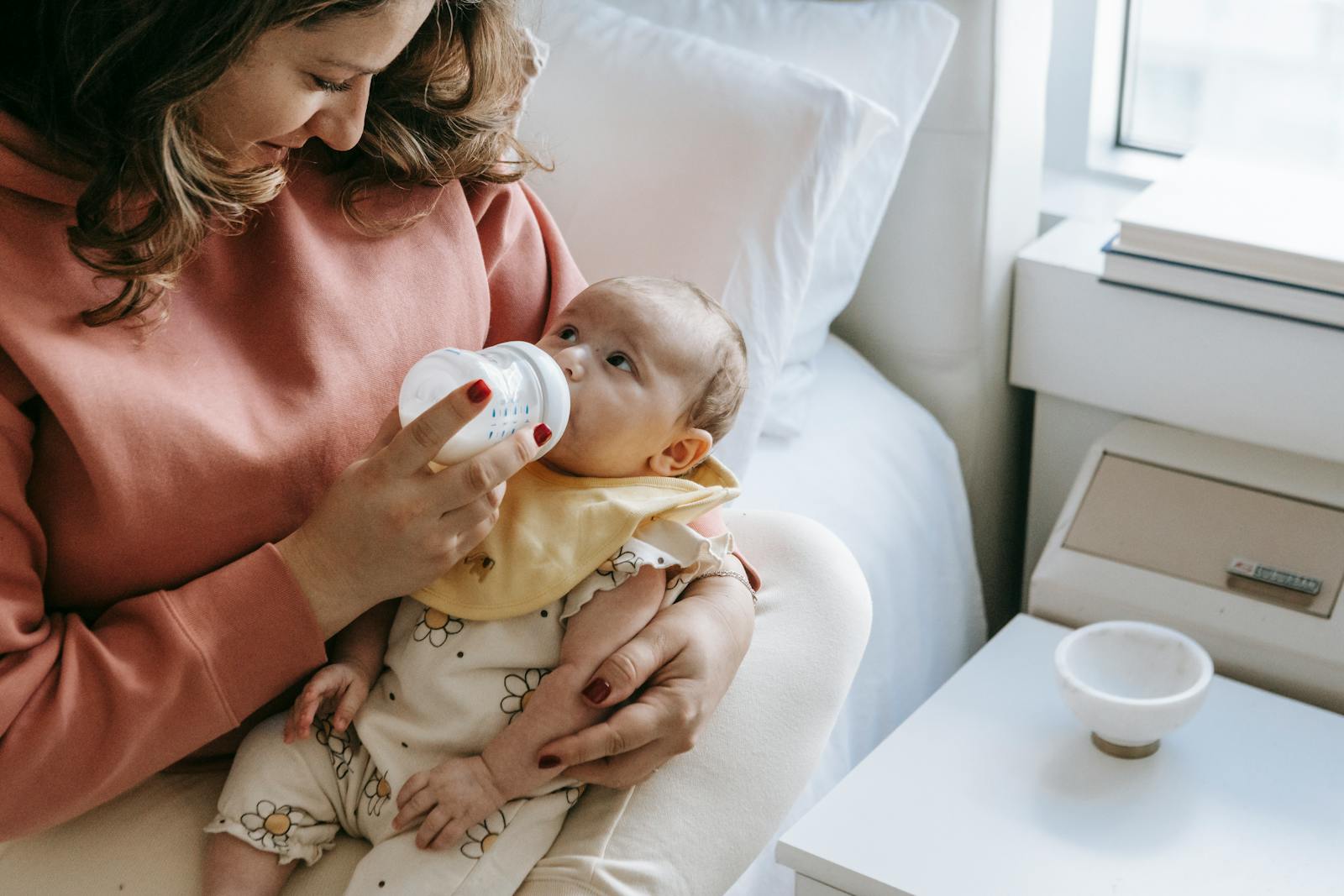 From above of crop happy young mother in casual clothes smiling while hugging and feeding adorable newborn from bottle sitting on comfortable bed
